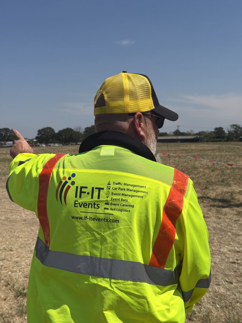 IF-IT Events marshal in a branded hi-vis jacket directing traffic in a field